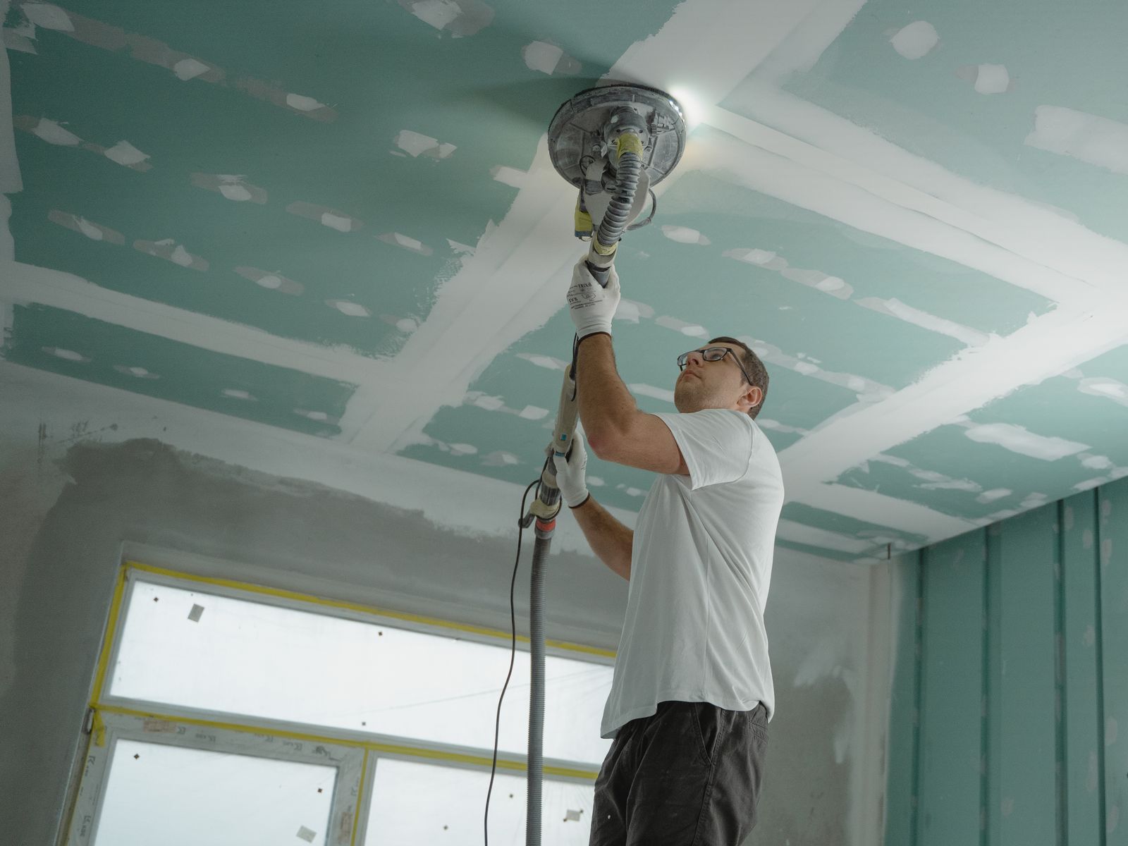 Tradesman using a dust-extracting sander to smooth a skim-coated drywall ceiling during a refinishing project