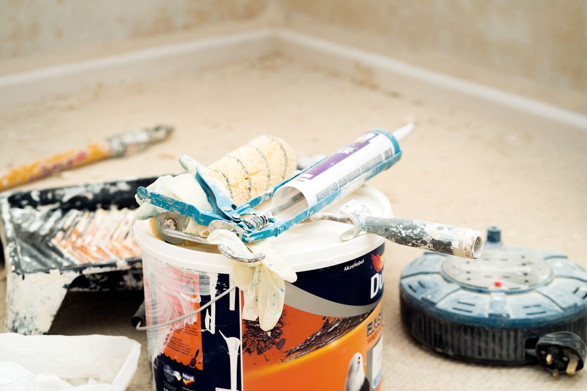 Paint bucket, roller sleeve, caulk gun, and taping tools staged on a protected floor
